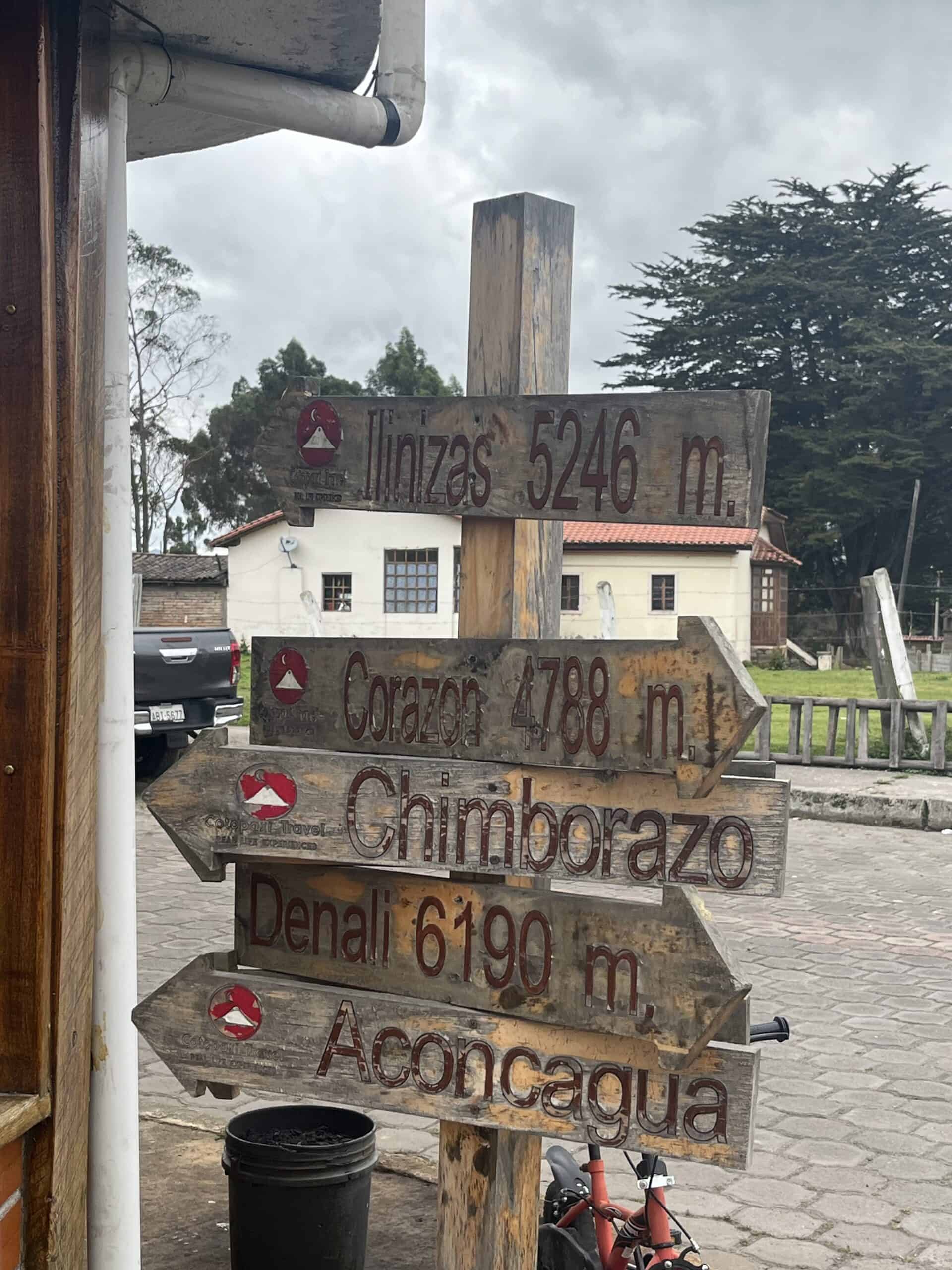 Signpost showing directions to various mountains, including Illinizas, Corazon, Denali, and Aconcagua.