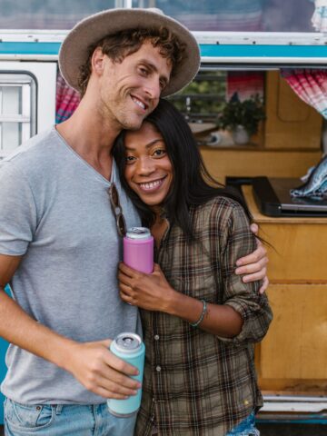 Couple holding drinks in custom koozies beside a vintage camper van during a road trip.