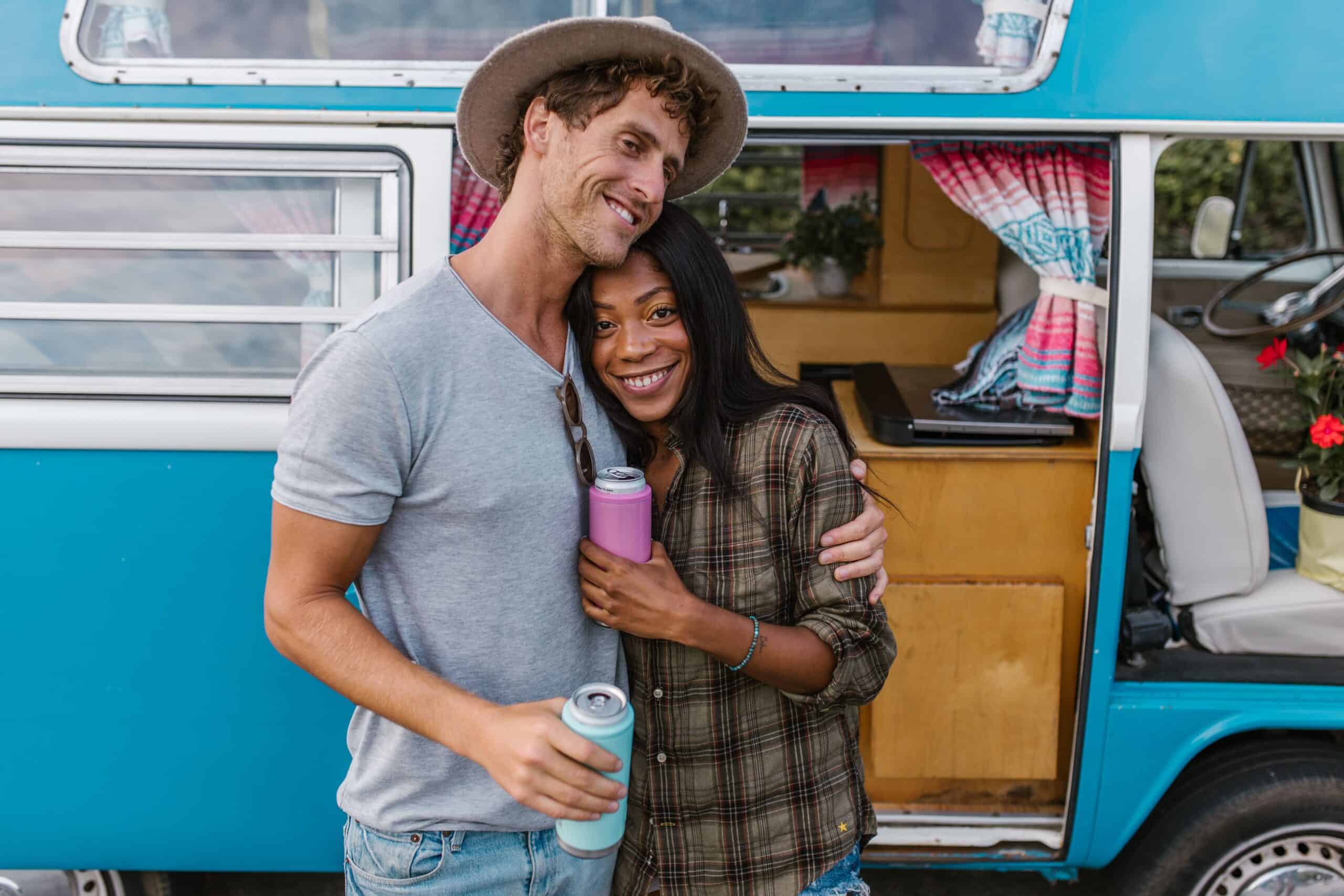 Couple holding drinks in custom koozies beside a vintage camper van during a road trip.
