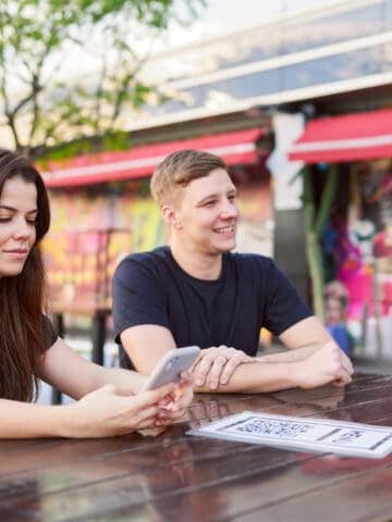 Travelers using a smartphone to access a QR code menu at an outdoor restaurant.