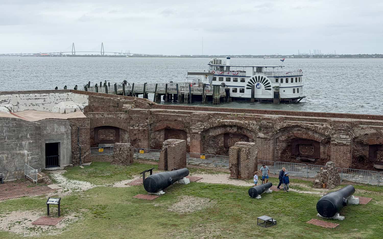 Cannons and brick ruins inside Fort Sumter with a tour boat docked beyond the wall.