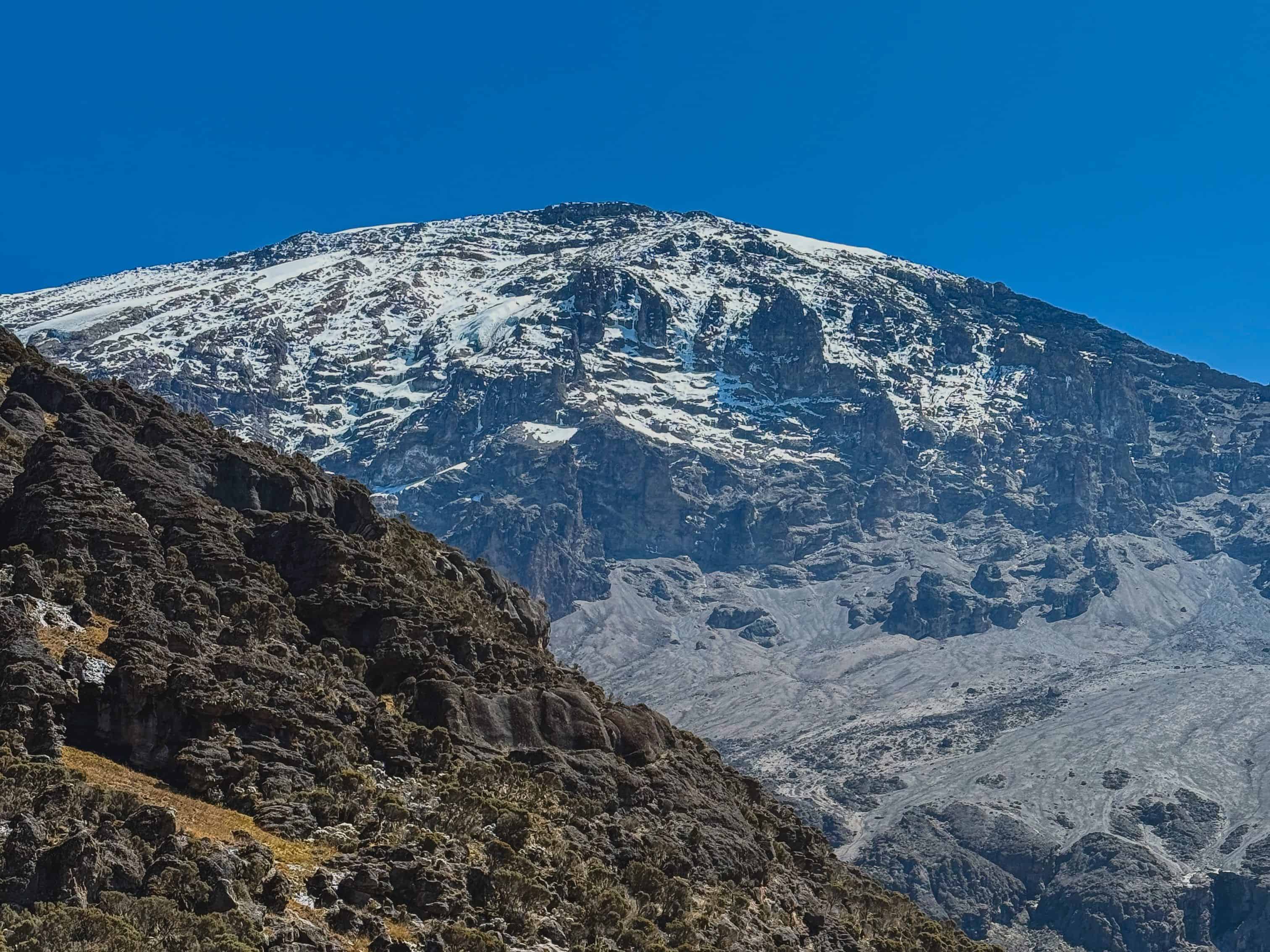 Snow-streaked cliffs on Kilimanjaro above a rocky foreground under a blue sky.