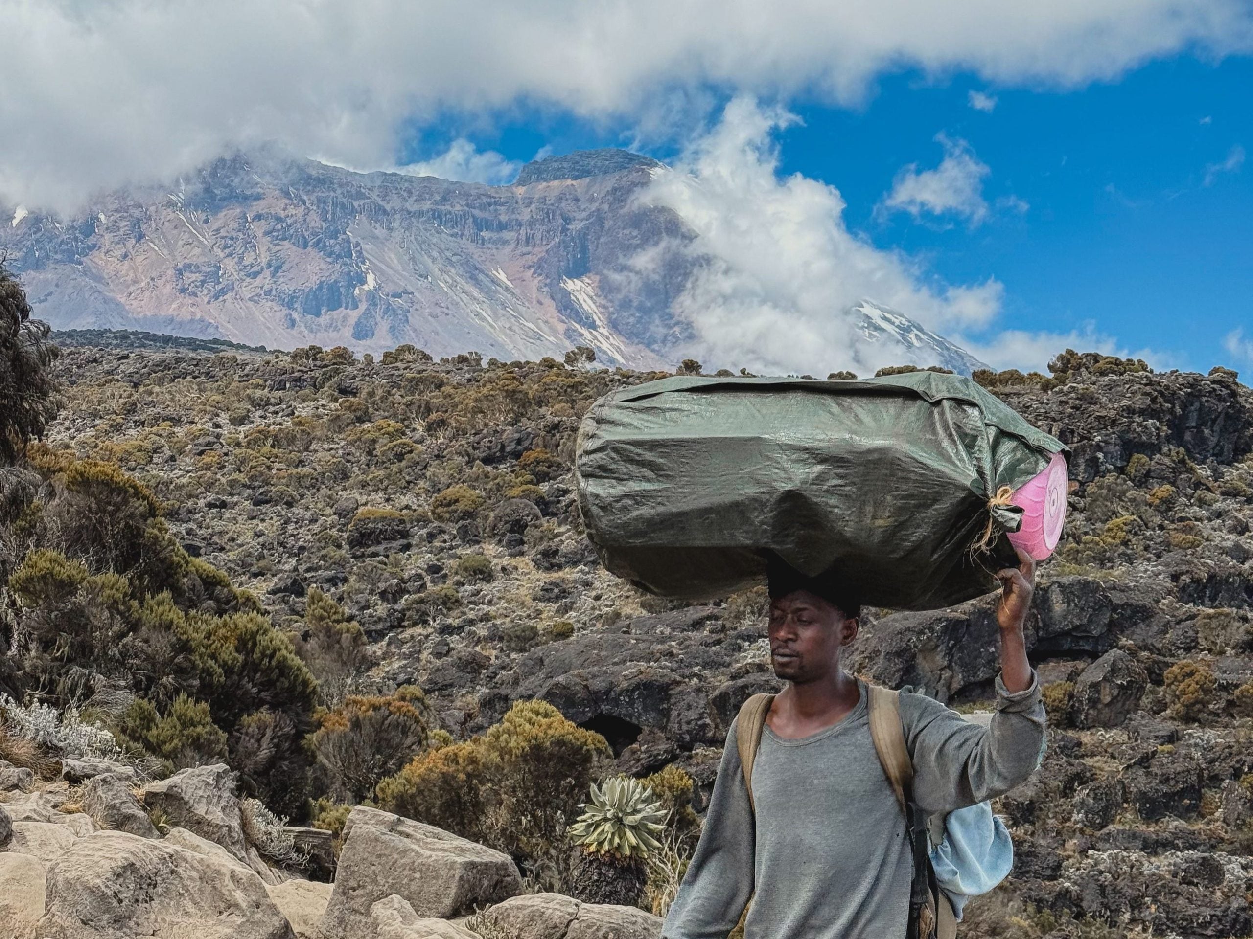 Porter carrying a large wrapped load on his head, with Kilimanjaro rising behind him.